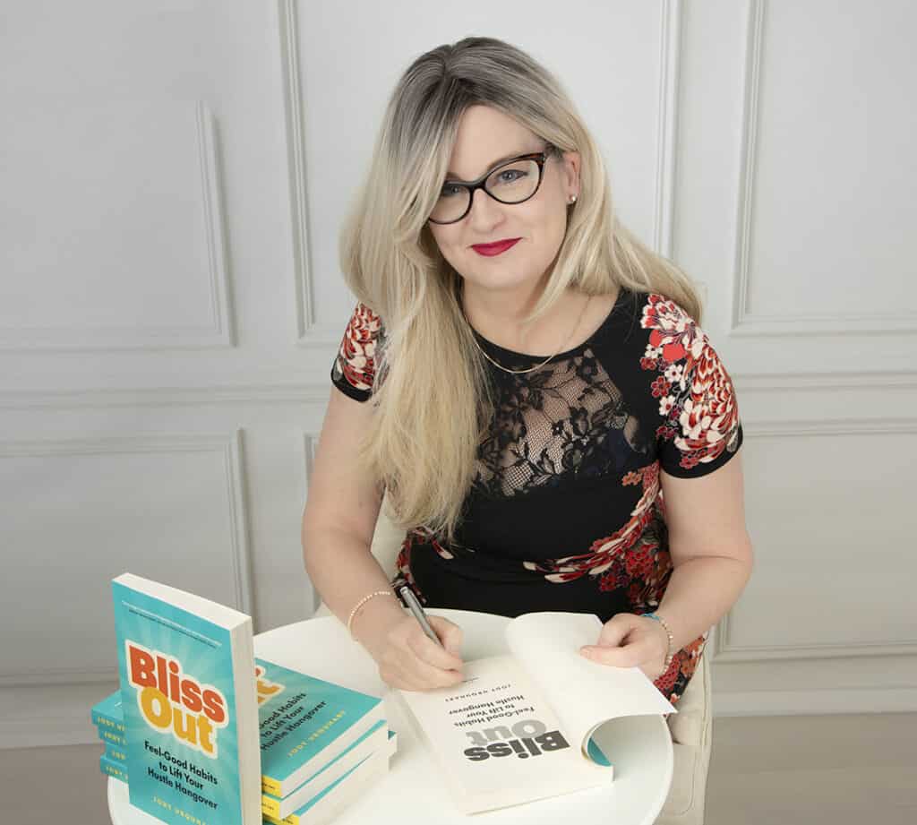 A woman with long blonde hair and glasses, wearing a black and floral dress, is smiling while signing her book "Bliss Out" at a table stacked with more copies, radiating powerful presence and vibrant communication. - Jodi O Photography
