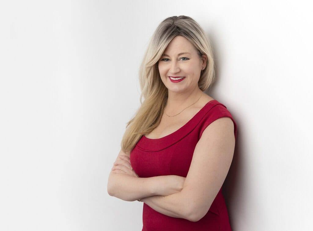A woman with long blonde hair, wearing a red dress, stands against a white background with her arms crossed and smiling, exuding a powerful presence. - Jodi O Photography