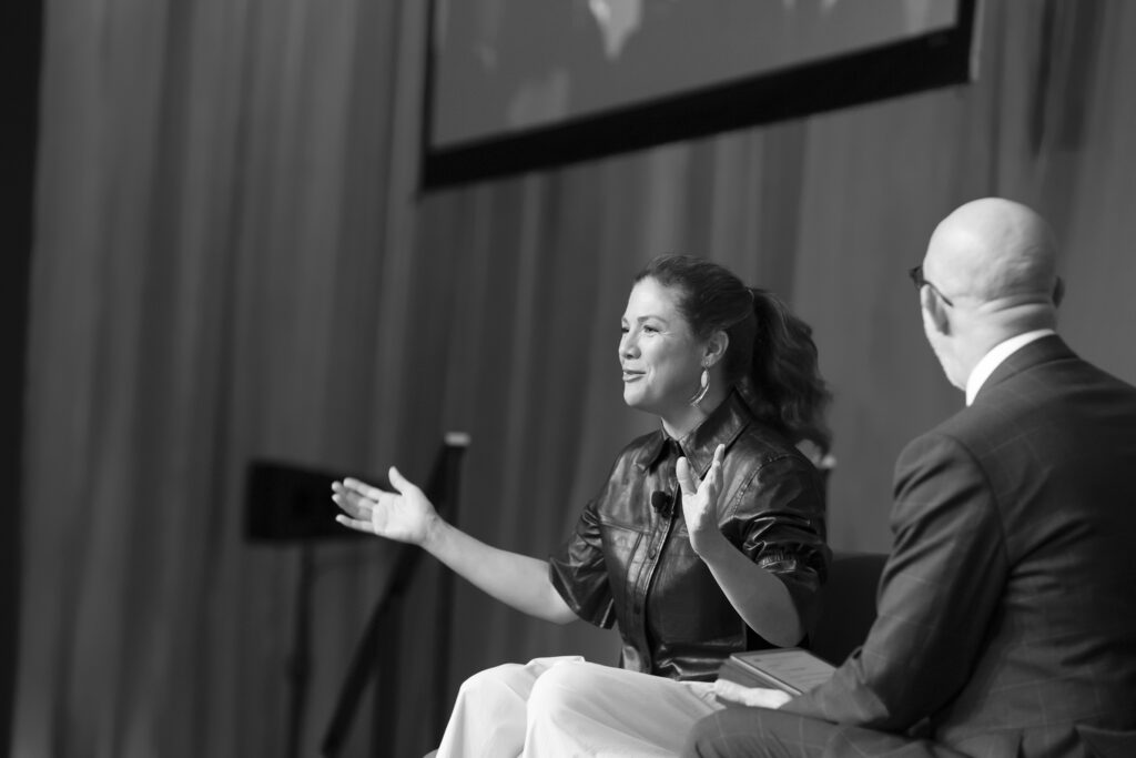 A woman gestures with open arms while speaking to a man in a suit during an on-stage conversation. Capturing speakers at professional events is essential—this black and white photo embodies the art of conference photography Calgary. - Jodi O Photography