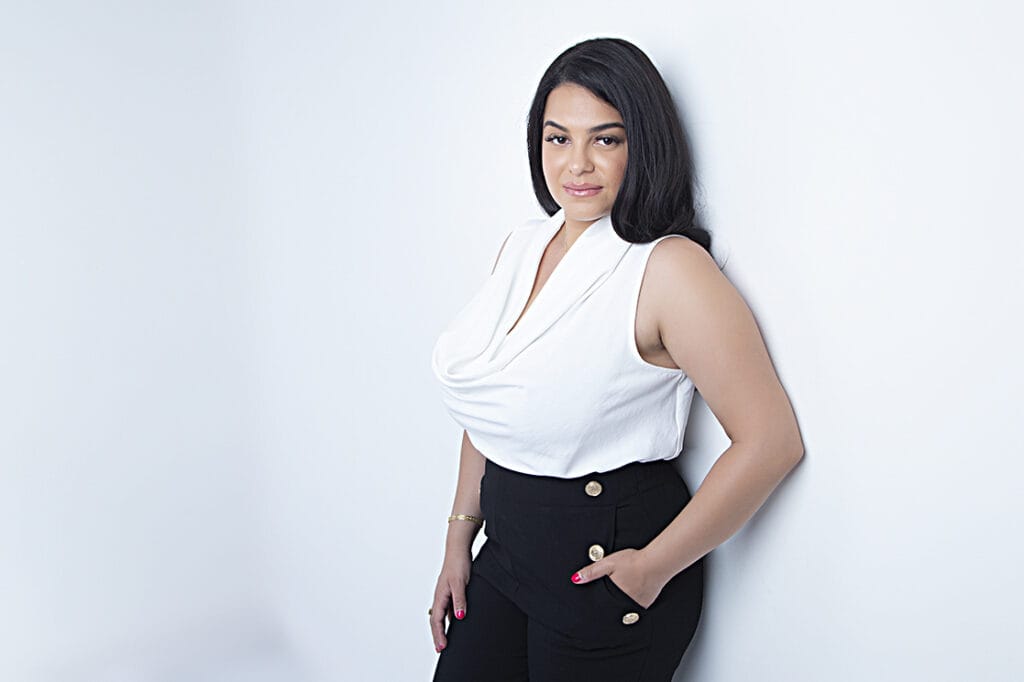 A woman with long dark hair stands against a white wall, wearing a sleeveless white top and high-waisted black pants with gold buttons—perfectly capturing confident headshots. She has one hand in her pocket and looks straight at the camera. - Jodi O Photography