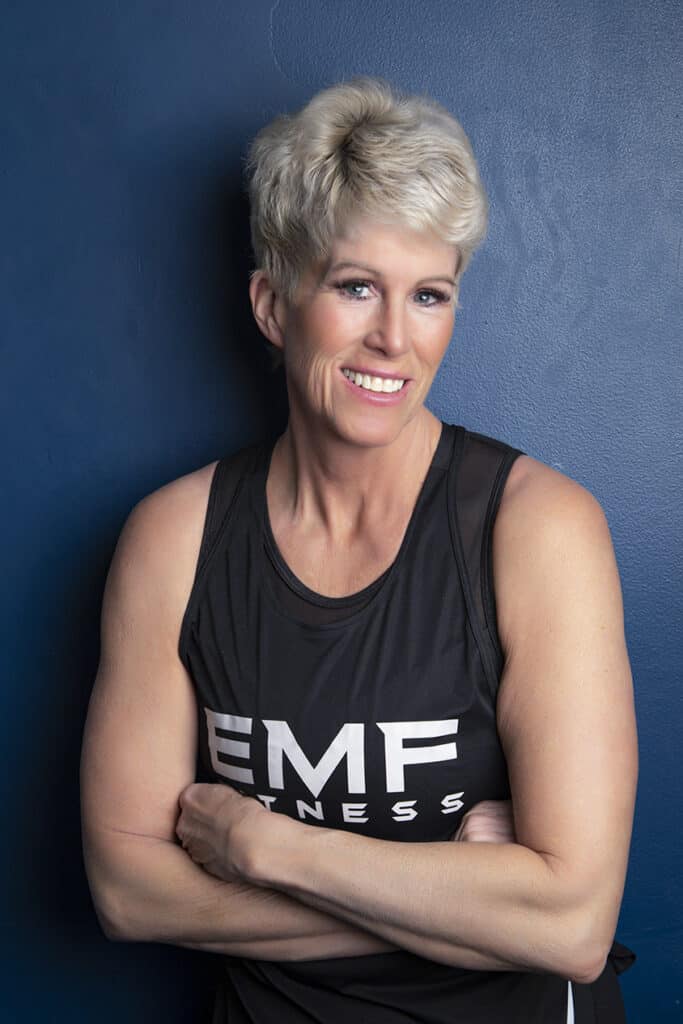 A smiling, fit woman with short blonde hair stands against a dark blue wall, arms crossed, wearing a sleeveless black top with “EMF FITNESS” in white letters—an excellent example of professional photography for modern headshots. - Jodi O Photography