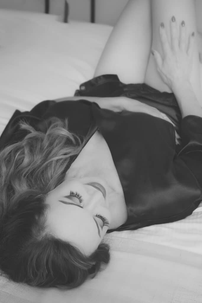A woman with long wavy hair lies on a bed, wearing a silky robe. She has her eyes closed and one hand resting on her thigh, appearing relaxed. The image is in black and white. - Jodi O Photography