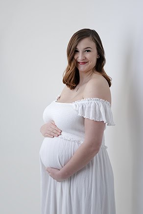 A pregnant woman with shoulder-length brown hair wearing an off-the-shoulder white dress smiles and cradles her belly, radiating joy in this elegant maternity post against a plain light background. - Jodi O Photography