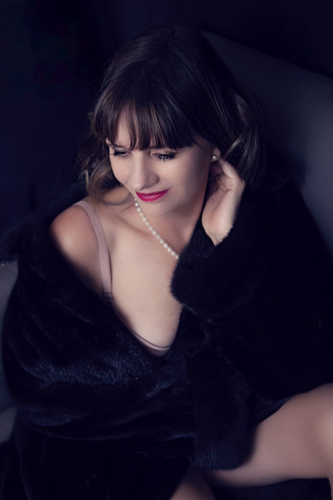 A woman with dark hair and bangs, wearing a pearl necklace, a black fur coat, and a light-colored top, sits and smiles softly while looking down, posed against a dark background. - Jodi O Photography