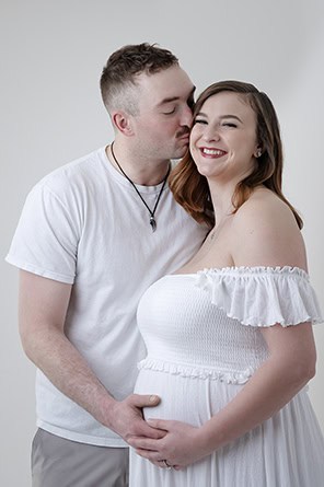 A man kisses a smiling pregnant woman on the cheek while standing behind her, both dressed in white. The man gently places his hand on her baby bump against a plain light background—perfect for a heartfelt Maternity Blog post. - Jodi O Photography
