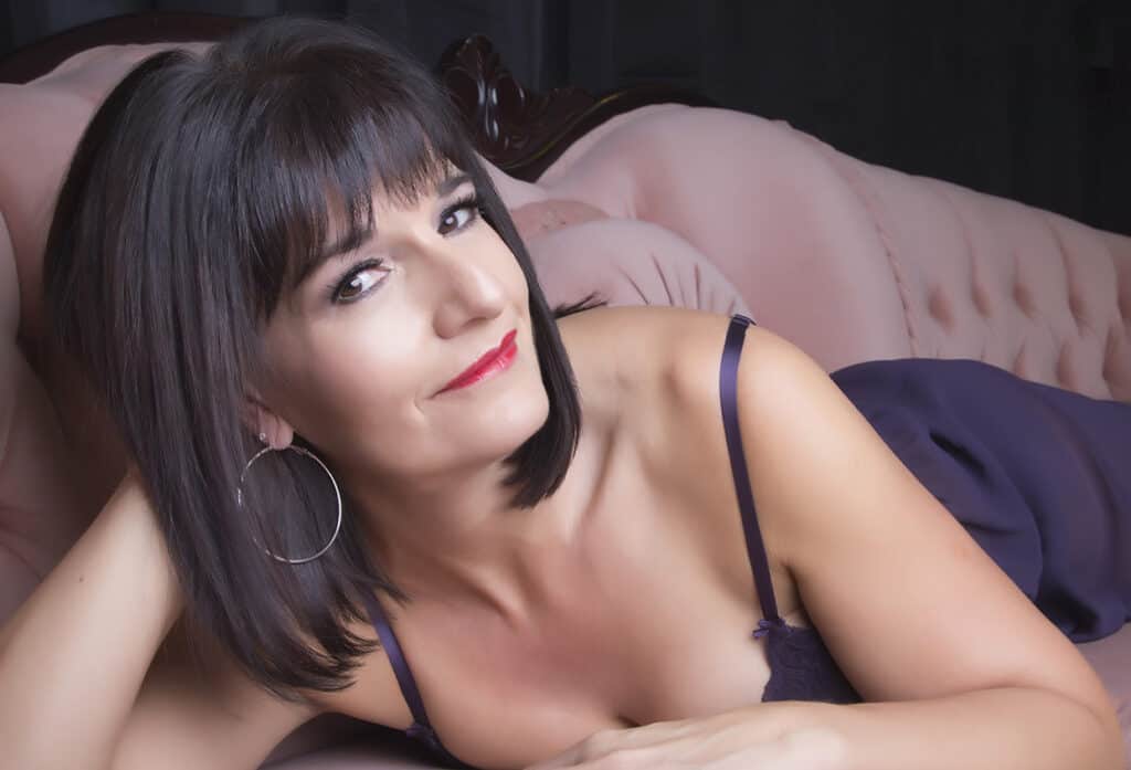 A woman with shoulder-length dark hair, wearing hoop earrings and a purple camisole, reclines on a pink tufted sofa, smiling slightly at the camera. - Jodi O Photography