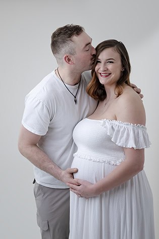 A man in a white shirt kisses a smiling pregnant woman on the forehead, gently placing his hands on her baby bump. The woman wears an off-the-shoulder white dress. This intimate maternity post is set against a plain light background. - Jodi O Photography