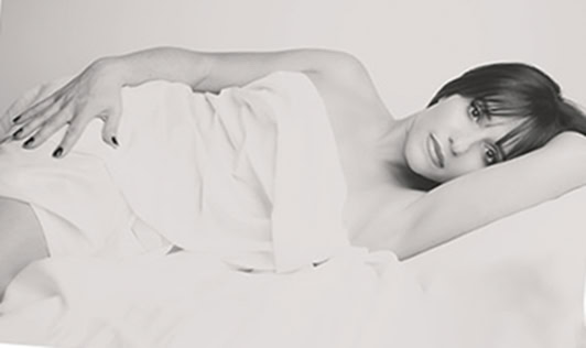 A woman with short dark hair lies on her side on a bed, covered in white sheets. She looks at the camera with a neutral expression, her left arm resting above her head and her right hand on the sheets. The image is in black and white. - Jodi O Photography