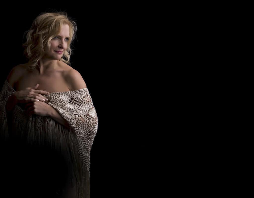 A woman with blonde hair looks to the side, smiling softly. She is draped in a crocheted shawl, exposing her shoulders, and stands against a black background with soft side lighting highlighting her face and upper body. - Jodi O Photography
