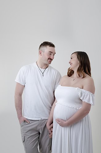 A pregnant woman in a white dress holds her belly and smiles at a man in a white t-shirt and beige pants. They stand close together, holding hands, celebrating their maternity journey against a plain light background. - Jodi O Photography