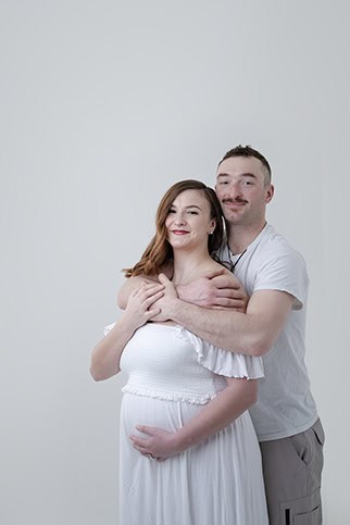 A smiling couple poses against a plain light background in this touching maternity moment. Perfect for a Maternity Blog, the woman cradles her baby bump while her partner stands behind her with arms lovingly wrapped around her shoulders. - Jodi O Photography