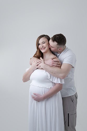 A man hugs and kisses a smiling pregnant woman from behind in this sweet maternity moment. Both dressed in white, the woman gently cradles her baby bump, standing against a plain light background. - Jodi O Photography
