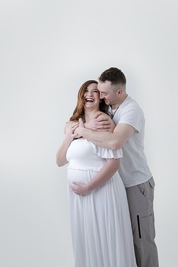 A smiling pregnant woman in a white dress stands in front of a man, who hugs her from behind. This joyful maternity moment is captured against a plain white background. - Jodi O Photography