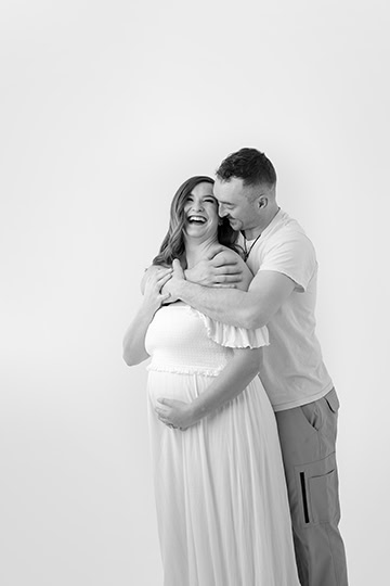 A smiling pregnant woman in a white dress stands in front of a man who hugs her from behind; both appear happy and relaxed, creating a heartwarming maternity post against a plain white background. - Jodi O Photography