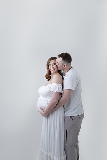 A smiling, pregnant woman in a white dress stands with a man behind her, embracing her from behind. Both are happy, wearing light-colored clothing, and beautifully capture the joy of pregnancy against a plain, light background. - Jodi O Photography
