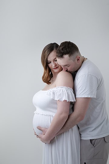 A man gently embraces a pregnant woman from behind in this Maternity Full Post, both dressed in white against a plain light background. She cradles her baby bump and smiles softly while he rests his head on her shoulder. - Jodi O Photography