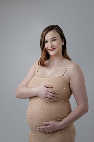 A pregnant woman in a beige, sparkly maternity dress smiles, with one hand resting on her belly and the other covering her chest, standing against a plain gray background. - Jodi O Photography