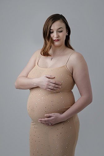 A pregnant woman in a fitted, nude-colored maternity dress with sparkles stands against a plain gray background, gently cradling her belly with one hand and resting her other hand on her chest. - Jodi O Photography