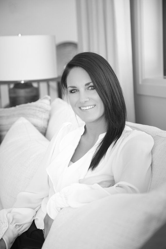 A woman with straight dark hair, dressed in a light blouse, sits on a couch and smiles at the camera in a well-lit room. This black and white portrait captures the essence of modern professionals, perfect for Calgary headshots. - Jodi O Photography