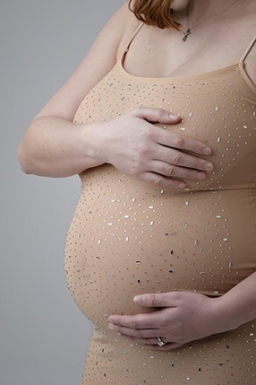 A pregnant woman in a sparkly, nude-colored dress cradles her belly with both hands, one above and one below, against a neutral background—perfect imagery for any Maternity Blog or Maternity Full Post. Her face is not visible. - Jodi O Photography