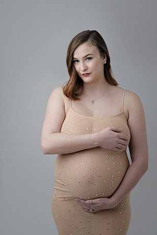 A pregnant woman in a beige, sparkly dress stands against a plain gray background, gently cradling her belly with one hand—a serene maternity post celebrating motherhood. She has light brown hair and a calm expression. - Jodi O Photography