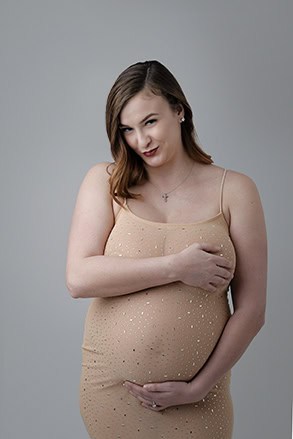 A pregnant woman in a beige, sleeveless dress with sparkling details poses for a graceful maternity post against a plain gray background, smiling slightly while cradling her belly and covering her chest with one hand. - Jodi O Photography