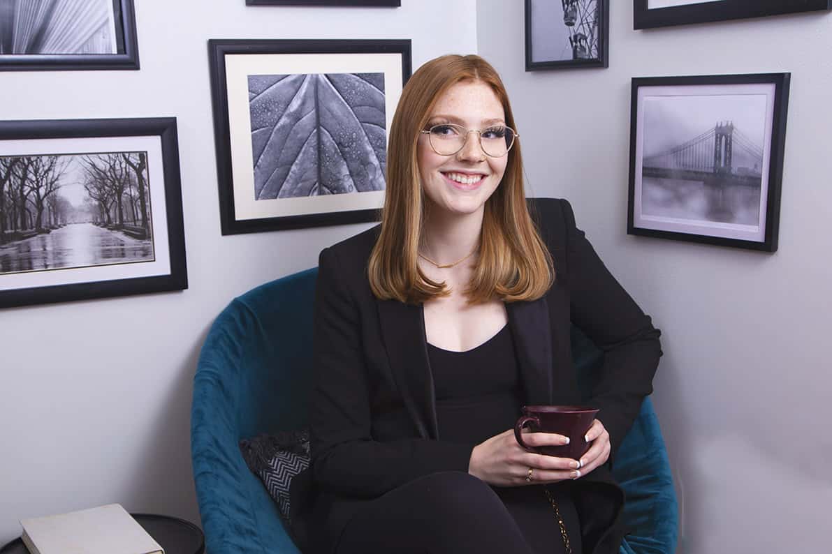 A woman with straight red hair and glasses sits smiling in a teal chair, holding a mug. Perfect for Calgary Headshots, she wears a black blazer, surrounded by black-and-white framed photos—a look modern professionals will love. - Jodi O Photography