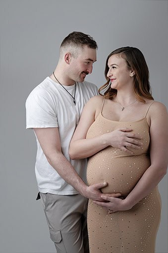 A smiling couple poses together in this beautiful maternity moment. The man stands behind the woman, wrapping his arms around her and resting his hands on her pregnant belly, perfectly capturing a loving Maternity Full Post. - Jodi O Photography