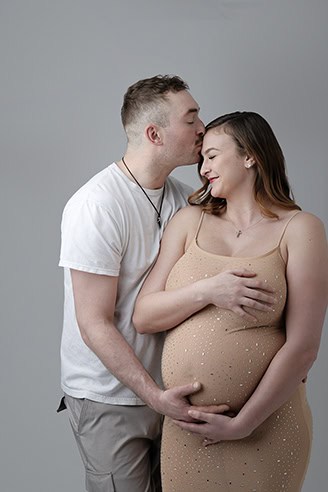A man in a white t-shirt kisses a smiling pregnant woman’s forehead. The woman, dressed in stylish maternity wear, holds her belly as the man gently cradles her baby bump. Both stand against a plain gray background. - Jodi O Photography