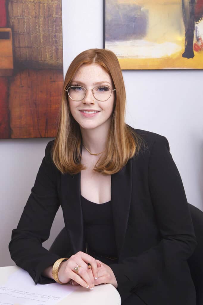 A young woman with straight, shoulder-length red hair and glasses sits at a desk, smiling at the camera. She wears a black blazer and holds a pen over some papers—an ideal example of Calgary Headshots for modern professionals. - Jodi O Photography