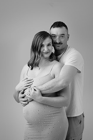 A man stands behind a pregnant woman, both smiling and embracing her belly in a joyful maternity post. The black and white photo features a plain background. - Jodi O Photography