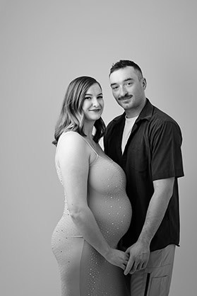 Black and white photo of a pregnant woman in maternity clothes, standing next to a man in a collared shirt. They are holding hands and smiling at the camera against a plain background, capturing a joyful moment in pregnancy. - Jodi O Photography