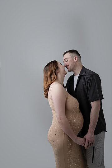A couple stands close together, smiling lovingly at each other. Celebrating her pregnancy, the woman wears a fitted, sleeveless beige dress and holds hands with the man, who is dressed in a black shirt and light-colored shorts. - Jodi O Photography