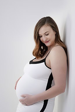 A pregnant woman with long brown hair, wearing a white and black dress, stands sideways and gently cradles her belly while smiling and looking down, creating a tender maternity moment against a plain white background. - Jodi O Photography