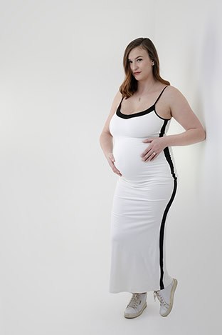 A pregnant woman in a fitted white maternity dress with black trim and white sneakers stands against a white wall, gently holding her belly and looking at the camera, radiating the beauty of pregnancy. - Jodi O Photography
