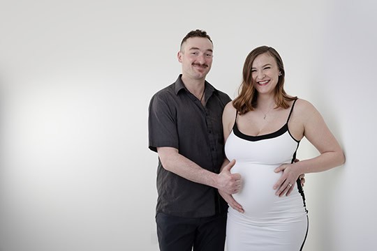 A smiling couple stands together against a plain white background. The woman, wearing a white dress, is visibly pregnant, and the man stands beside her with his hands gently on her belly—a beautiful moment perfect for a Maternity Blog. - Jodi O Photography