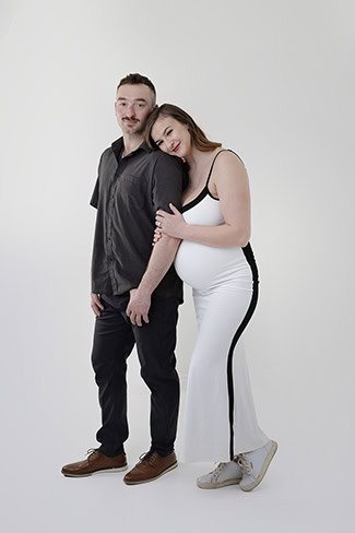 A maternity moment: A pregnant woman in a white dress leans affectionately on her partner’s shoulder. Both are smiling softly, creating a tender scene against a plain white background—perfect for sharing as a Full Post. - Jodi O Photography