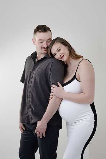 A pregnant woman in a white dress stands smiling and leaning on a man in a dark shirt, who looks down with a gentle expression. This intimate maternity moment is beautifully captured against a plain light background. - Jodi O Photography