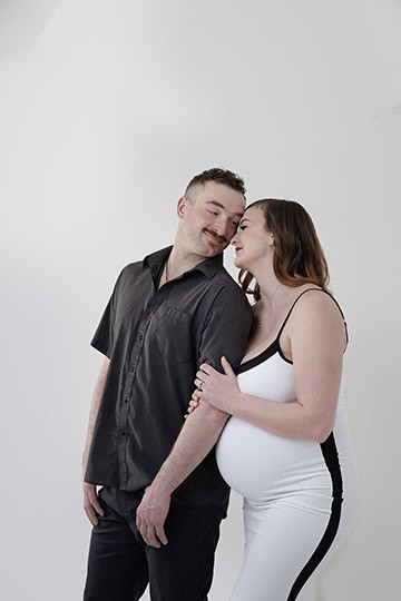 A maternity photo shows a pregnant woman in a white dress with black accents leaning affectionately on a man's shoulder. Both smile softly against a plain white background. See the full post for more tender moments. - Jodi O Photography