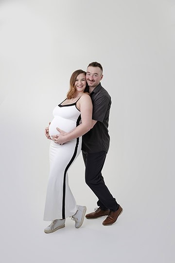 A smiling pregnant woman in a white dress and sneakers stands beside a man in a dark shirt and pants. The man hugs her from behind, his hands on her belly, capturing a joyful maternity full post against a plain white background. - Jodi O Photography