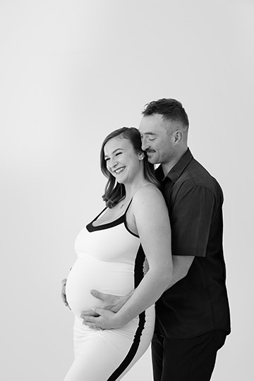 A smiling pregnant woman in a sleeveless dress stands in front of a man who gently holds her belly from behind. Both are happy and looking slightly to the side in this black and white maternity post. - Jodi O Photography