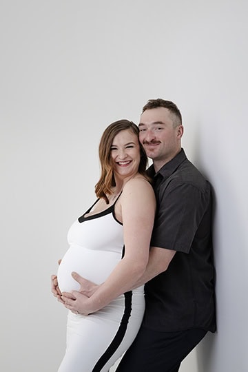 A smiling couple poses against a white wall in this charming Maternity Full Post. The woman, wearing a white dress, is visibly pregnant as her partner in a dark shirt lovingly cradles her baby bump from behind. - Jodi O Photography