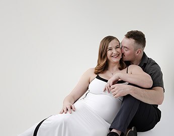 A smiling pregnant woman in a white dress sits beside a man in dark clothing who is kissing her cheek. This joyful maternity post captures their closeness as they sit together against a plain white background. - Jodi O Photography