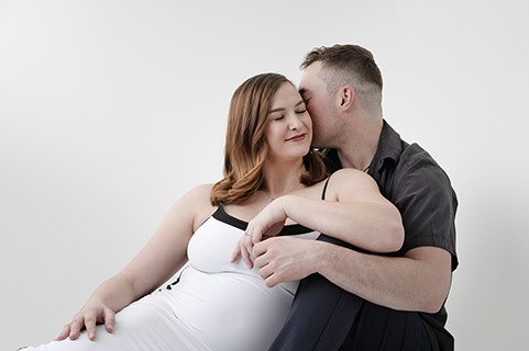 A man lovingly kisses a smiling, pregnant woman on the cheek in this heartwarming maternity post. They sit close together against a plain white background, with her in a white dress and him in a dark shirt. - Jodi O Photography
