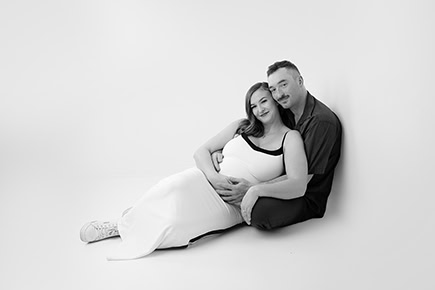 A couple sits on the floor against a white wall in this black and white Maternity Full Post. The woman, in a flowing white dress, leans into her partner as he lovingly rests his hand on her pregnant belly. Both are smiling gently. - Jodi O Photography