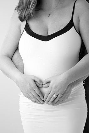 A pregnant woman in a sleeveless dress stands with a man's arms wrapped around her, both forming a heart shape with their hands on her belly. This black-and-white image is perfect for a heartfelt maternity blog or maternity post. - Jodi O Photography