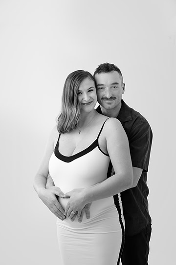 A smiling couple poses in a studio. The woman, wearing a fitted dress, stands in front while the man stands behind her with his hands gently forming a heart over her pregnant belly, capturing a joyful maternity moment. - Jodi O Photography