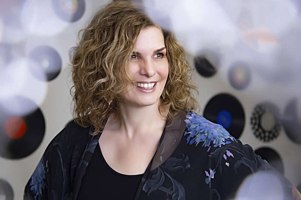 A woman with curly blonde hair smiles while looking to the side. Perfect for Calgary headshots, her dark floral-patterned top adds flair, and the background features blurred vinyl records on a wall—a stylish choice for modern professionals. - Jodi O Photography
