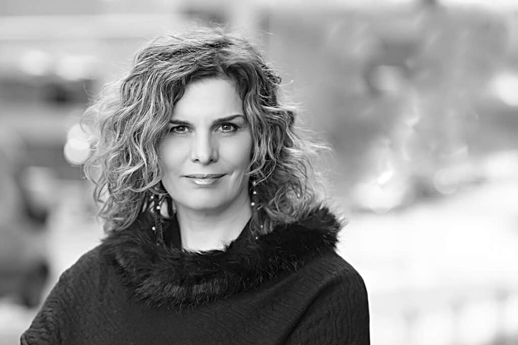 Black and white portrait of a woman with wavy, shoulder-length hair, wearing a dark top with a fur collar—ideal for Modern Professionals or Calgary Headshots. She looks directly at the camera with a neutral expression against a softly blurred background. - Jodi O Photography