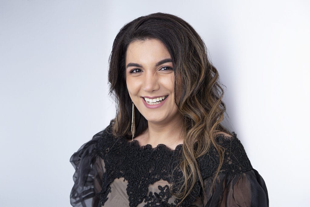 A woman with wavy brown hair and a black lace top smiles at the camera against a plain white background, capturing the confident essence of Modern Professionals in Calgary Headshots. - Jodi O Photography